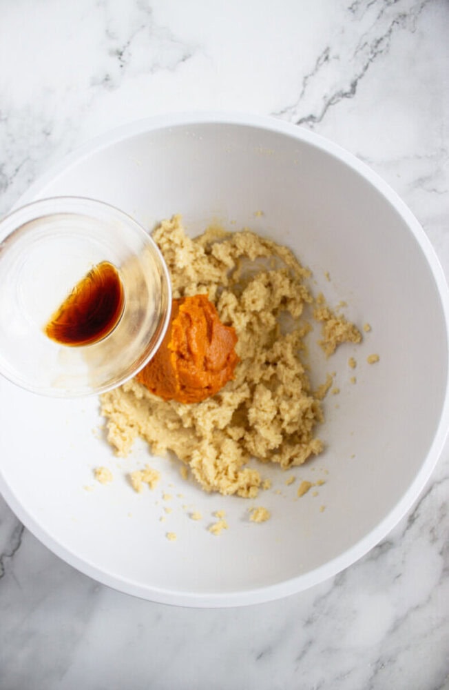A glass bowl of vanilla extract being added to a white mixing bowl containing creamed butter, sugar, and pumpkin puree on a marble countertop.