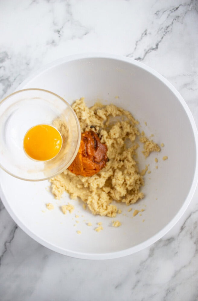 A glass bowl with a cracked egg is being poured into a white mixing bowl containing cookie dough and pumpkin puree on a marble countertop.