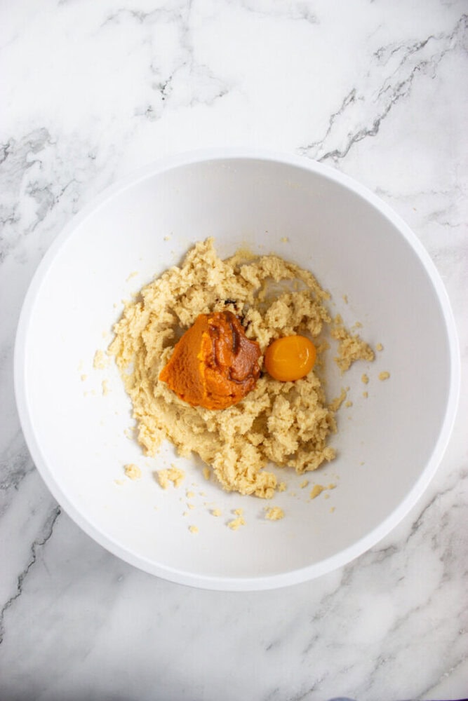 A white mixing bowl containing creamed butter and sugar, with a scoop of pumpkin puree and an egg on top, on a marble countertop.
