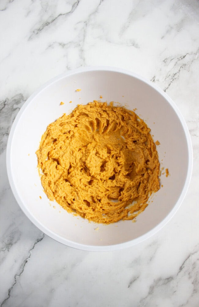 A white bowl containing mixed orange cookie dough sits on a white marble countertop.