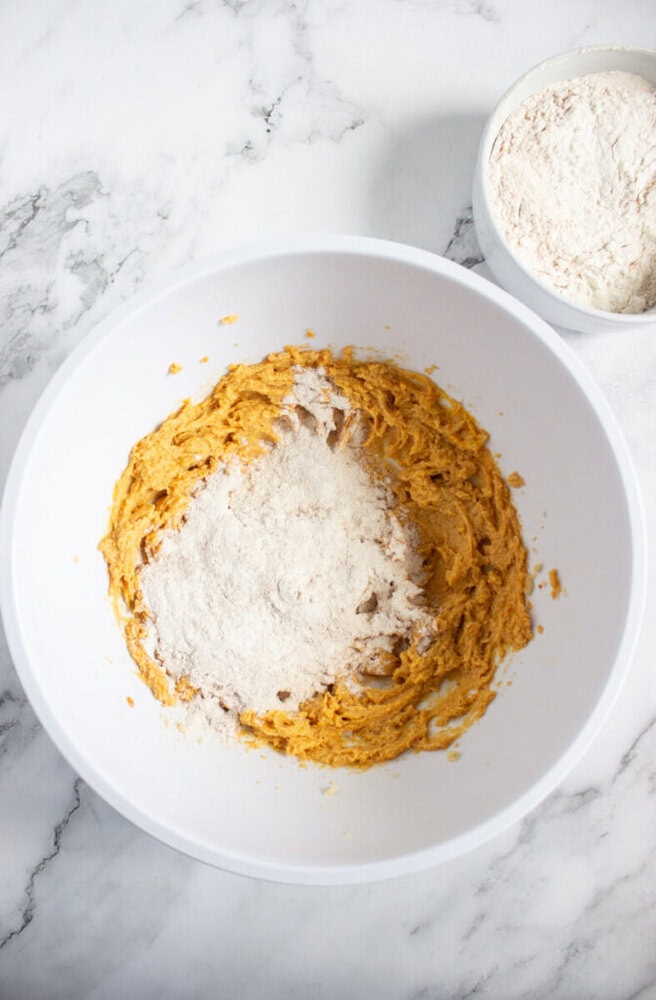 A white mixing bowl with partially mixed pumpkin batter and flour on top, next to a small bowl of flour, on a marble countertop.