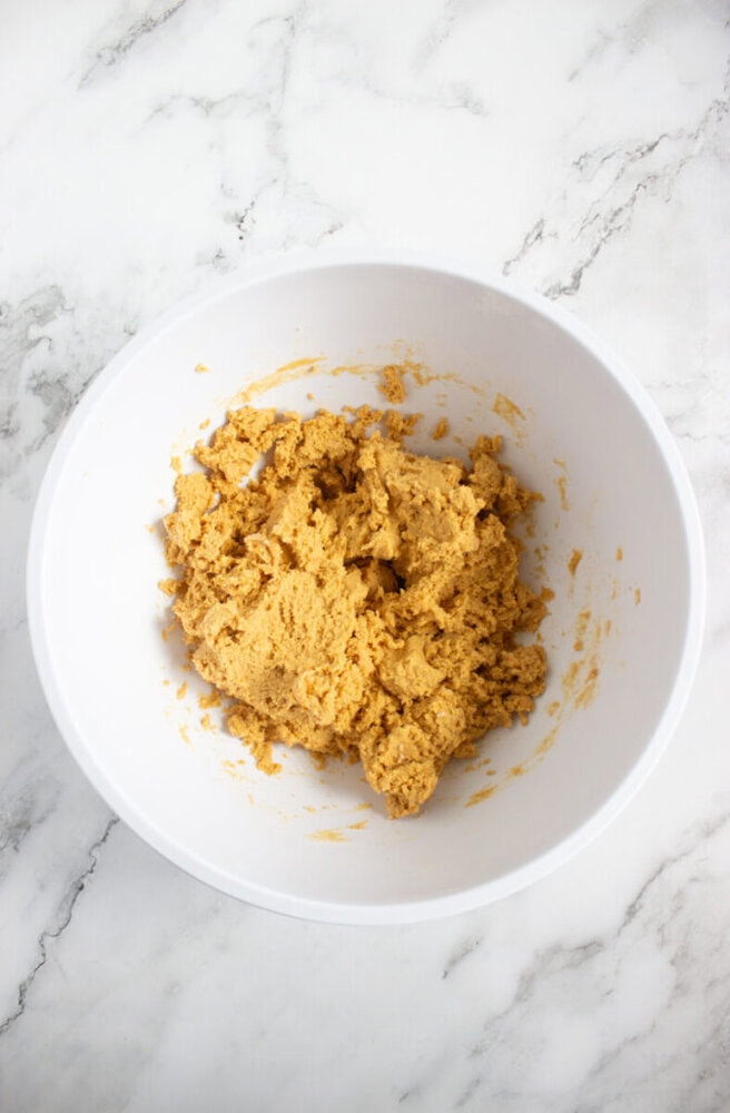 A white mixing bowl containing a portion of unbaked cookie dough sits on a white marble surface.