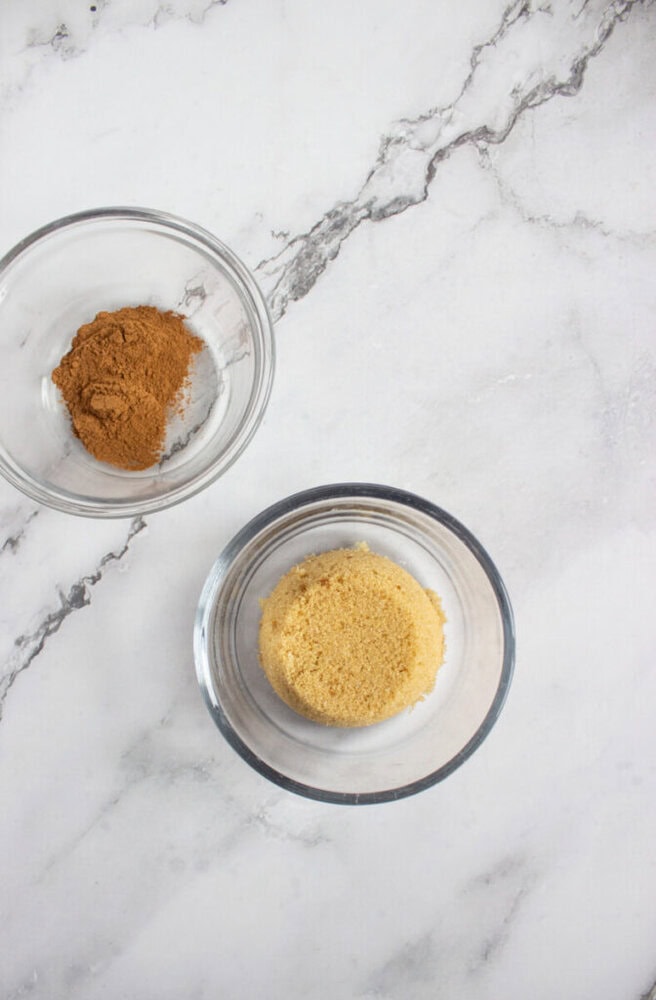 Two small glass bowls on a marble surface, one containing ground cinnamon and the other filled with light brown sugar.