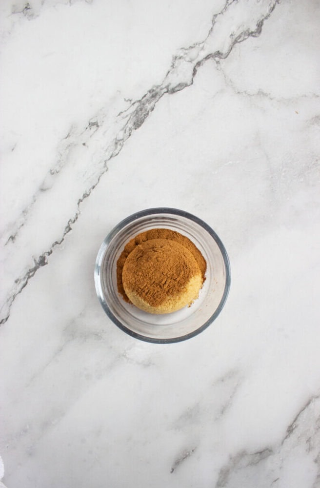 A small glass bowl containing a mixture of ground cinnamon and sugar on a white marble surface.