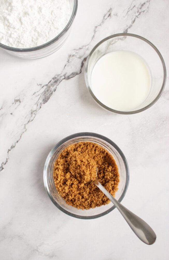 Three glass bowls on a marble surface: one with flour, one with milk, and one with brown sugar and a spoon.