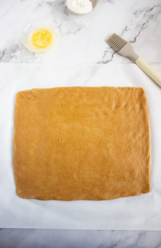 A rectangular sheet of dough on parchment paper, with melted butter, flour, and a pastry brush nearby on a marble surface.