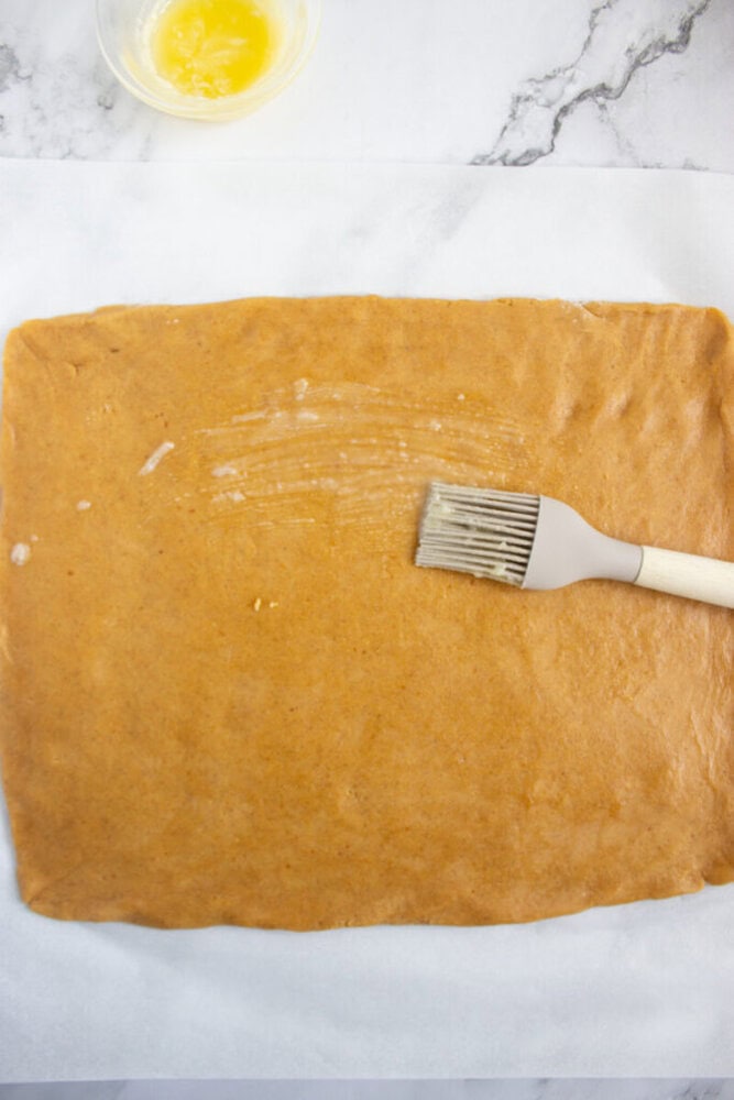 A rectangular sheet of dough on parchment paper is being brushed with melted butter using a pastry brush. A small bowl of melted butter sits nearby.