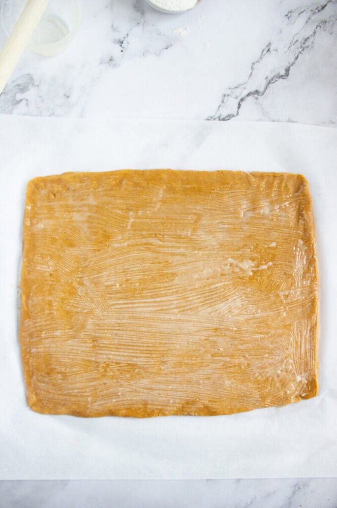 A rectangular sheet of dough is rolled out on parchment paper, with visible brush marks on its surface, on a marble countertop.