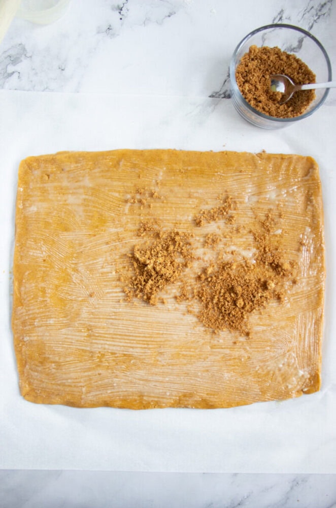 A rectangular sheet of dough spread with butter, topped with piles of brown sugar, sits on parchment paper next to a bowl of brown sugar with a spoon.