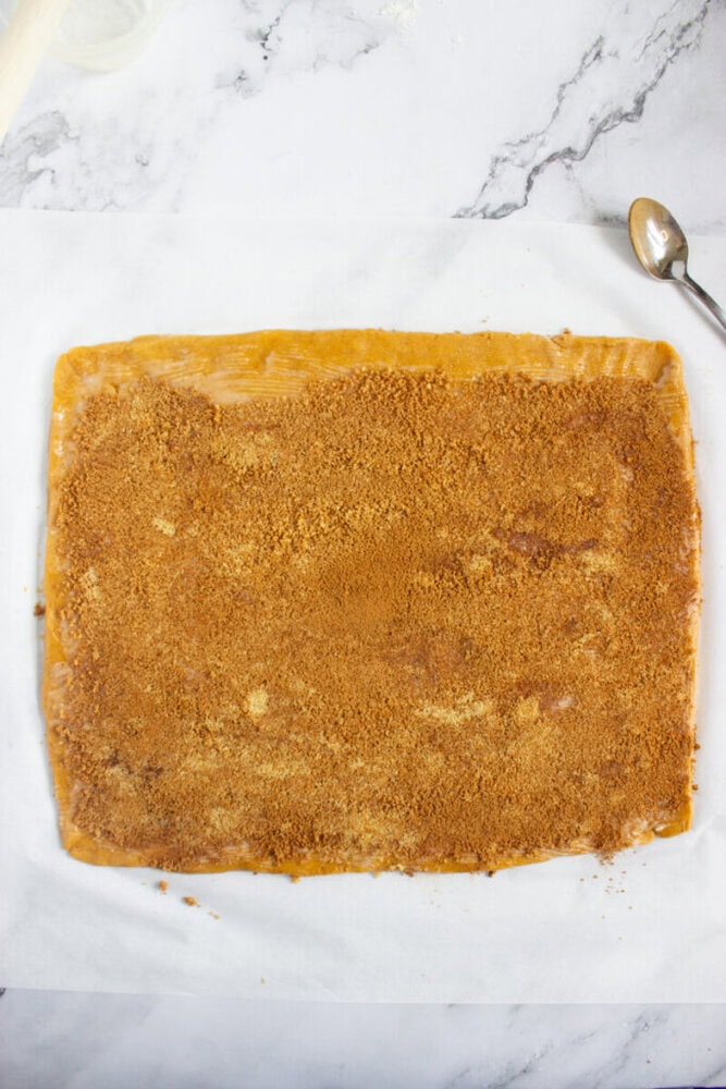 A rectangular sheet of dough topped evenly with a layer of brown sugar and cinnamon, placed on parchment paper next to a metal spoon.
