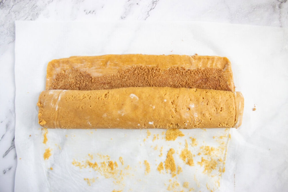 Rolled dough with cinnamon sugar filling being prepared on parchment paper atop a marble surface.