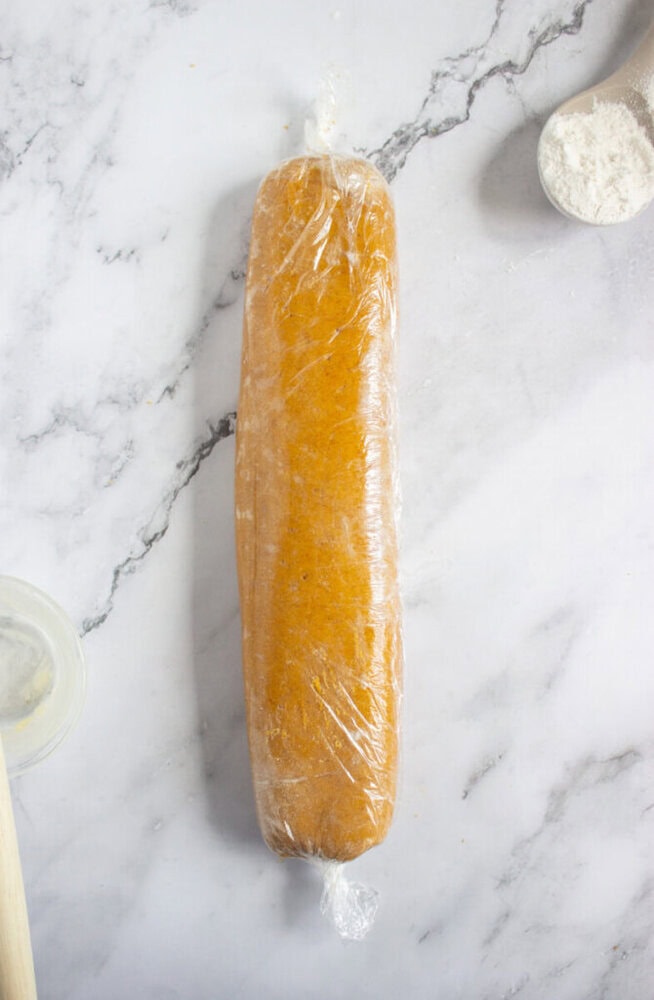 A log of cookie dough wrapped tightly in plastic wrap rests on a marble countertop near a wooden spoon and a bowl of flour.