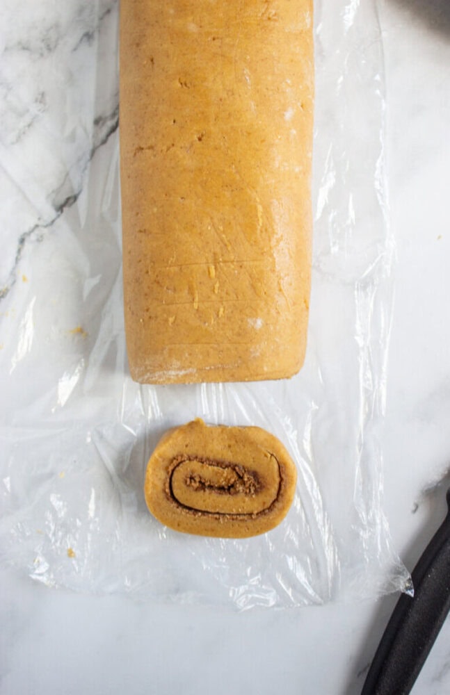 A log of rolled dough with a visible spiral cut slice sits on a piece of plastic wrap on a white marble surface.