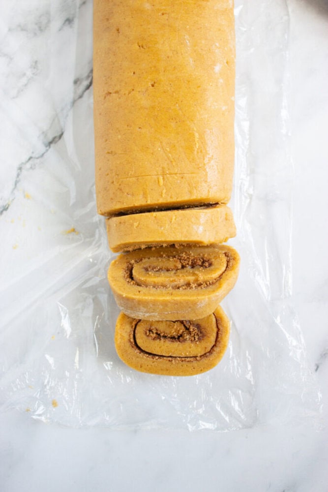 A rolled log of dough with a cinnamon filling is partially sliced on a sheet of plastic wrap, showing spiral layers inside each slice.