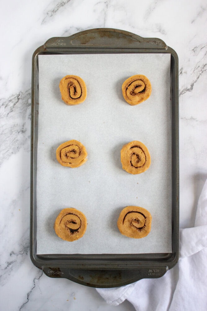 A baking tray lined with parchment paper holds six evenly spaced unbaked cinnamon rolls on a marble surface.