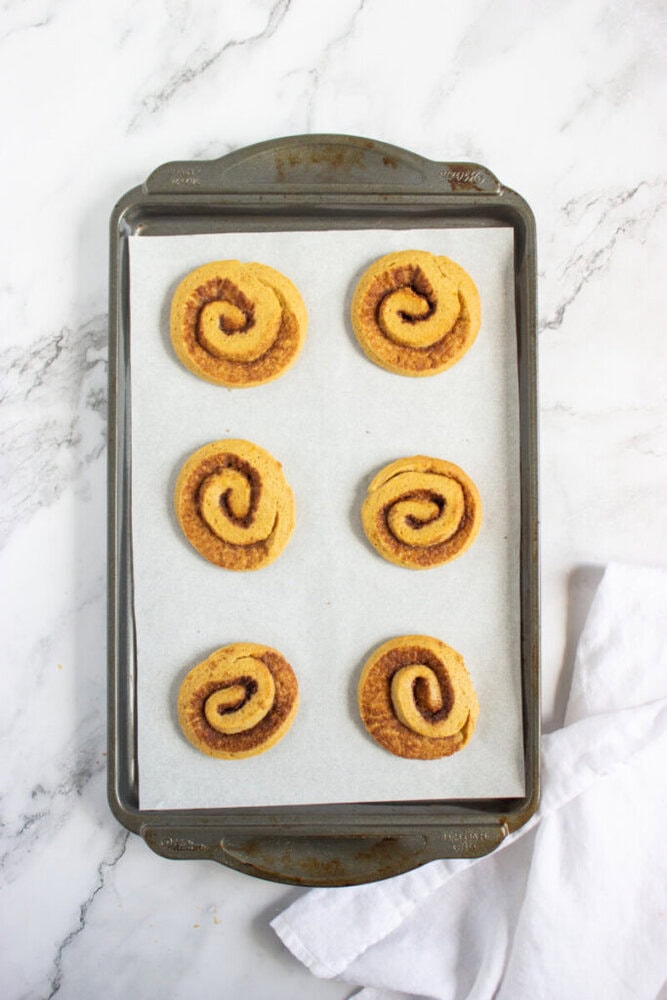A baking tray lined with parchment paper holds six baked cinnamon rolls arranged in two rows on a marble surface, with a white cloth nearby.