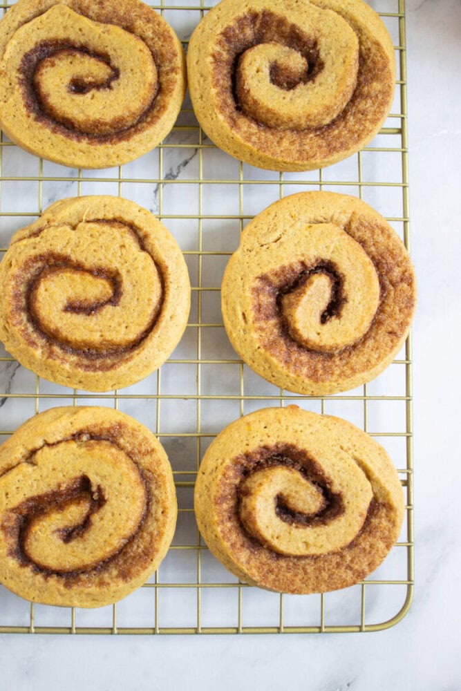 Six cinnamon rolls on a cooling rack, viewed from above on a white marble surface.