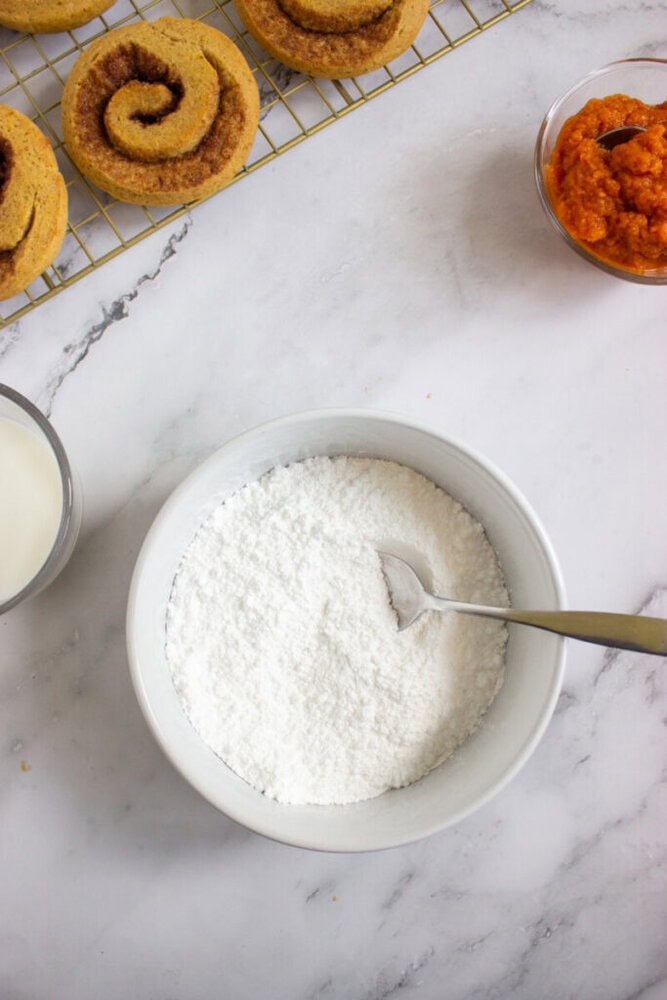 A bowl of powdered sugar with a spoon, next to a glass of milk, a bowl of orange puree, and cinnamon rolls on a cooling rack.