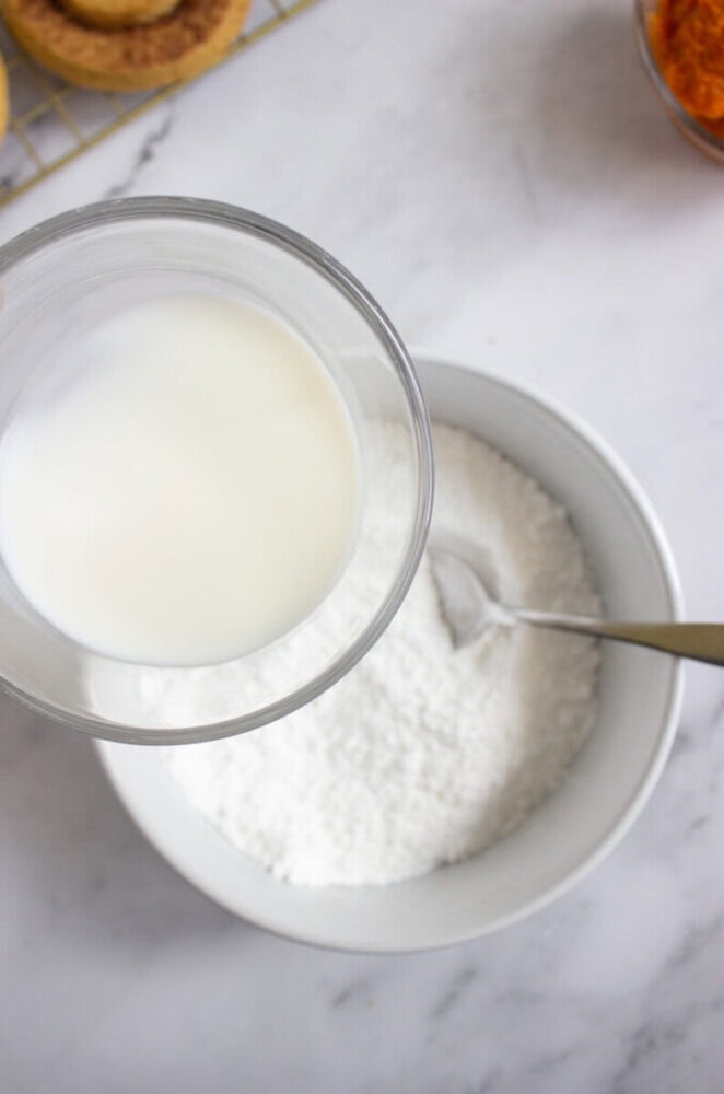 A glass container of milk held above a white bowl filled with powdered sugar and a spoon on a marble countertop.