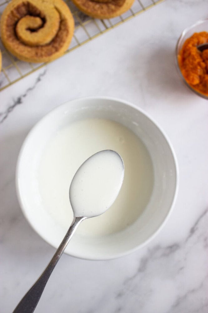 A spoon holding white icing above a bowl, with a cooling rack of cinnamon rolls and a small bowl of orange puree nearby on a marble surface.