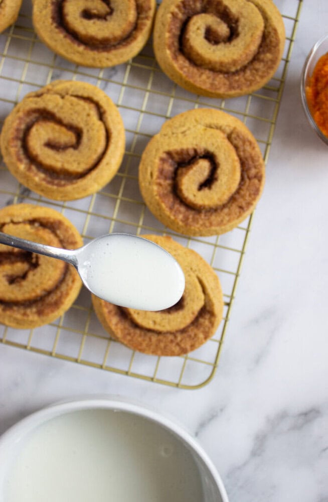 A spoon with white icing is held above pumpkin cinnamon rolls cooling on a wire rack, with a bowl of icing and a small dish of pumpkin nearby.
