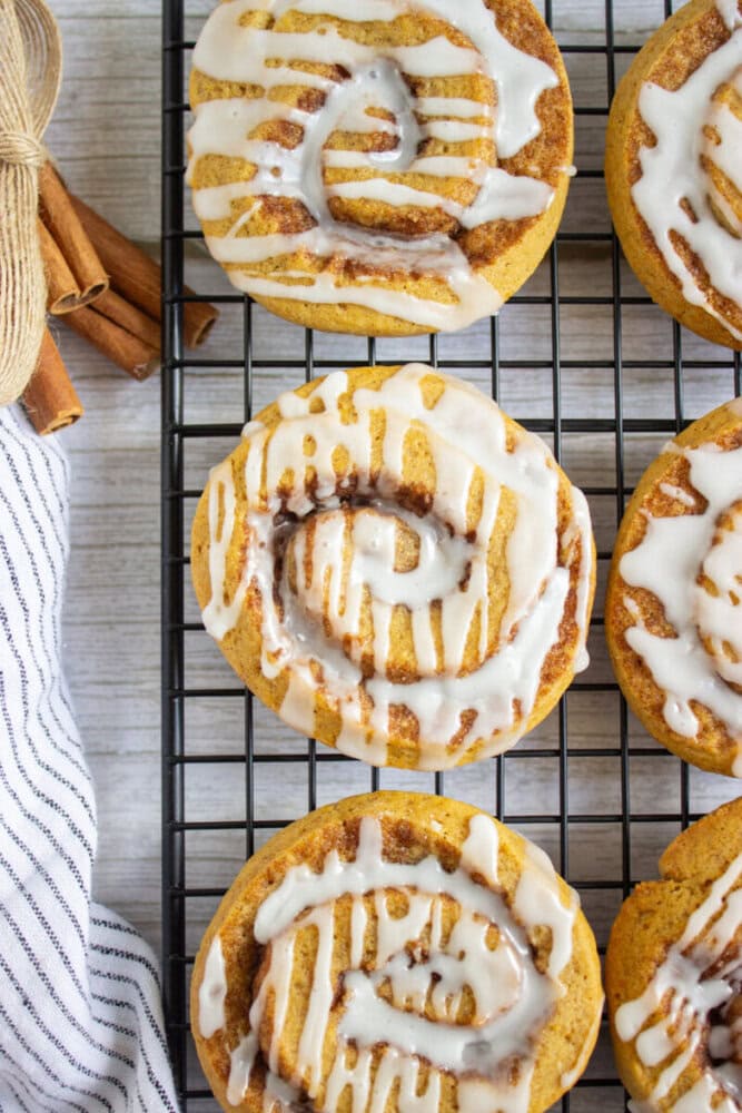 Iced cinnamon rolls arranged on a cooling rack with a striped towel and cinnamon sticks visible to the side.