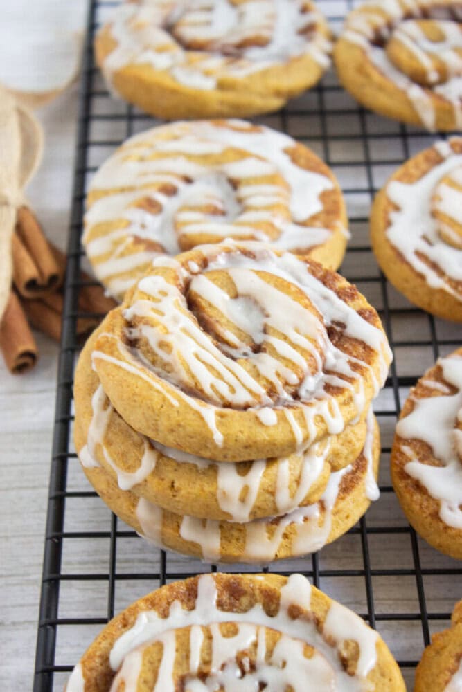 Three stacked iced cinnamon rolls and several others cooling on a black wire rack, with a drizzle of white icing on each roll.