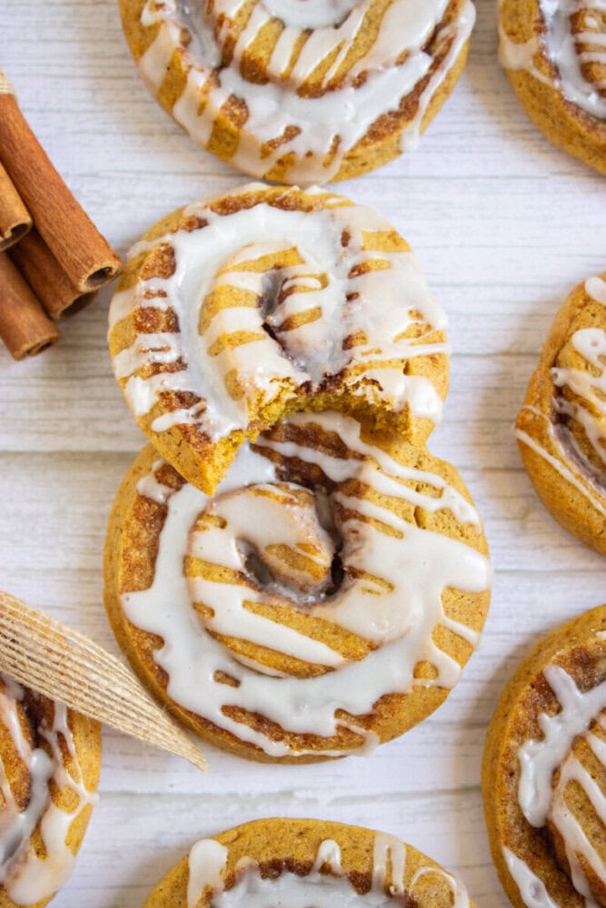 Iced cinnamon rolls are arranged on a light wooden surface, with one partially eaten roll on top and cinnamon sticks placed nearby.