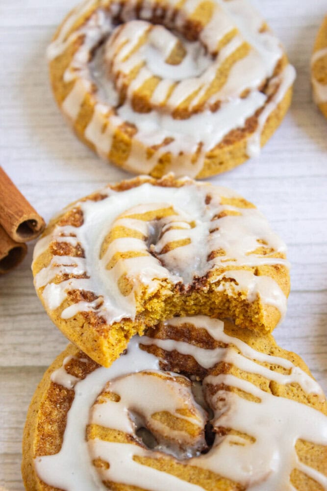 Close-up of three iced cinnamon rolls on a white surface, with one roll partially bitten and two cinnamon sticks visible on the side.