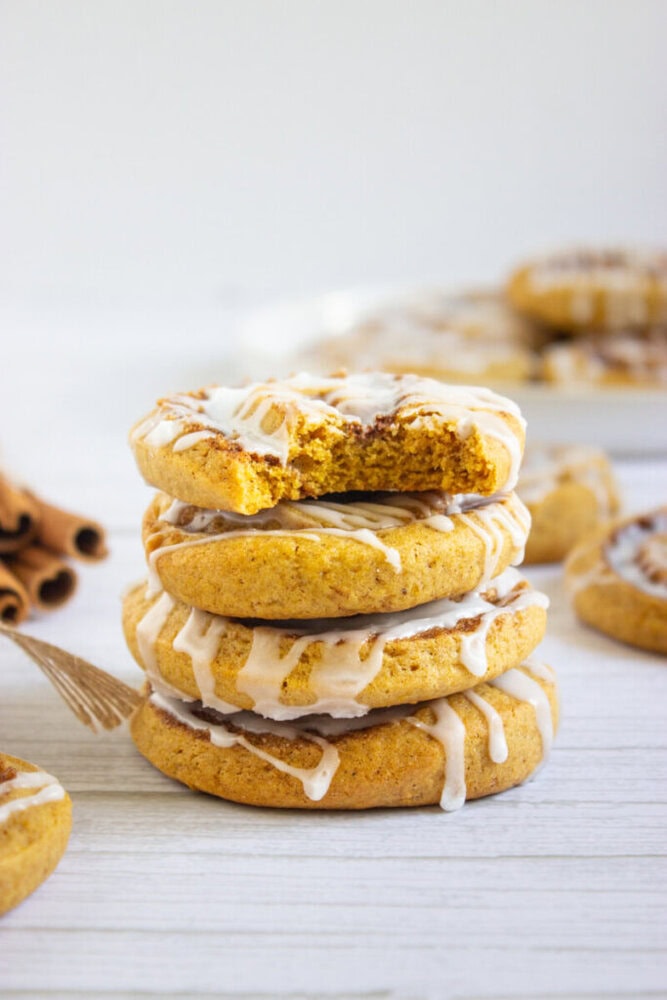 A stack of four glazed cookies, with the top cookie missing a bite, sits on a light wooden surface. Additional cookies and cinnamon sticks are in the background.