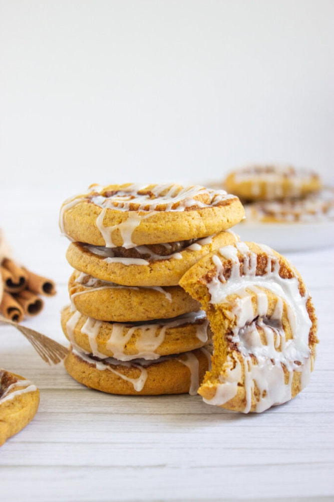 A stack of glazed cookies with a bite taken out of the top one, placed on a white wooden surface with cinnamon sticks in the background.