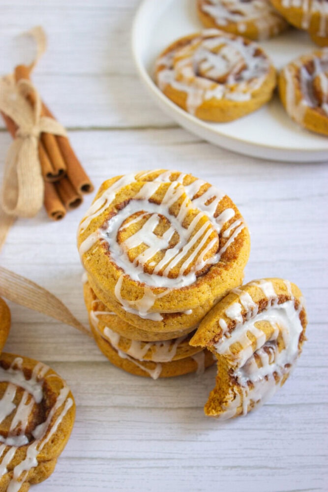 Three pumpkin cinnamon rolls drizzled with icing are stacked on a table, with a bite taken from the top roll. More rolls and cinnamon sticks are visible in the background.