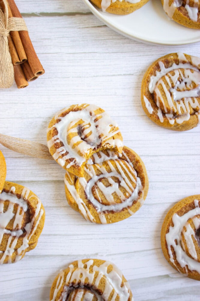 Iced cinnamon swirl cookies arranged on a white surface, with a plate of cookies and a bundle of cinnamon sticks in the background.