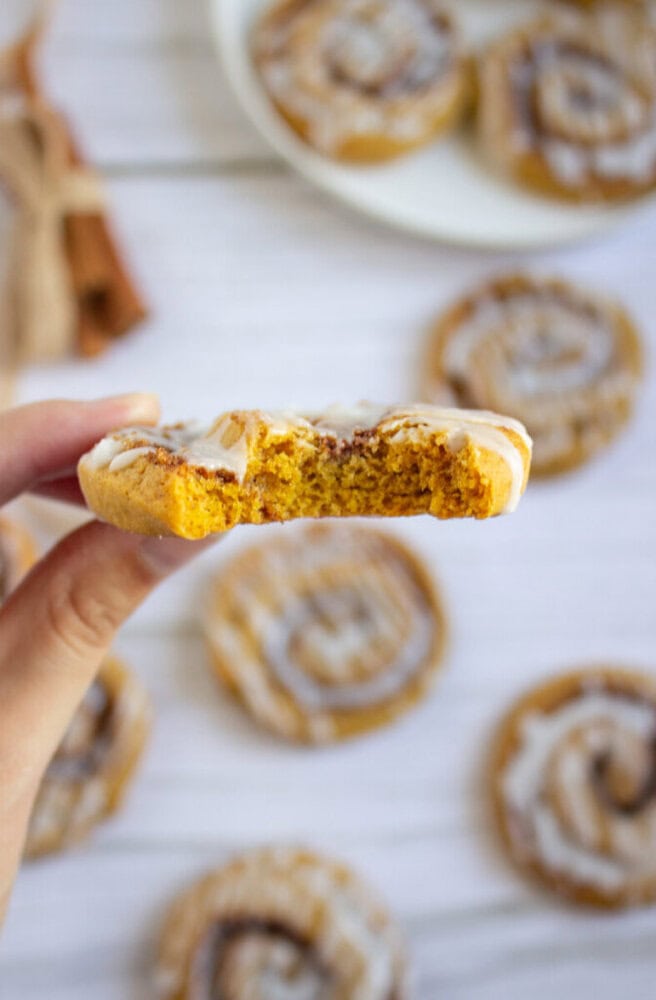 A hand holds a bitten iced cookie with a cinnamon swirl; other similar cookies are scattered on a white surface in the background.