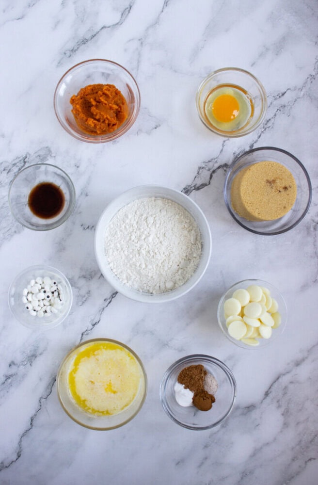 Various baking ingredients in glass bowls arranged on a marble surface, including flour, egg, sugar, butter, pumpkin puree, vanilla, chocolate chips, and spices.