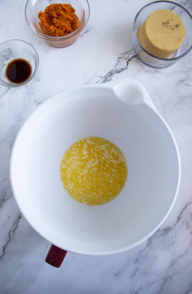 A white mixing bowl with melted butter sits on a marble surface, surrounded by small bowls containing pumpkin puree, brown sugar, and vanilla extract.