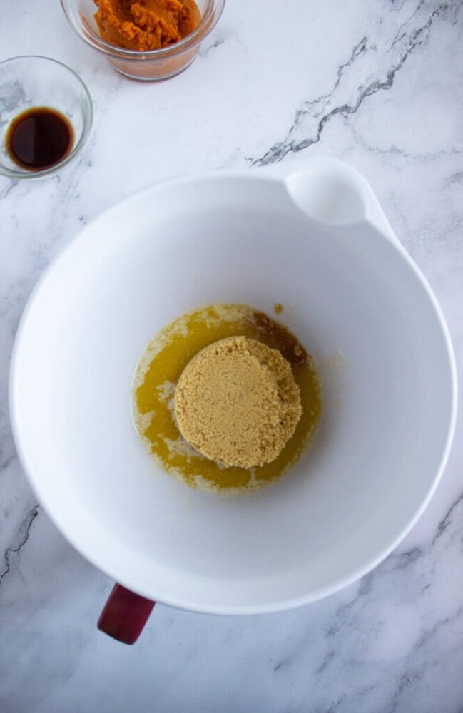 A white mixing bowl with melted butter and brown sugar, with small bowls of vanilla extract and pumpkin puree nearby on a marble countertop.