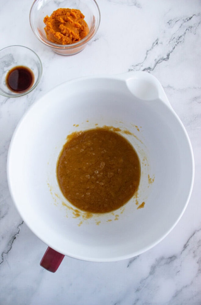 A mixing bowl with a brown liquid mixture, next to a small bowl of vanilla extract and a bowl of pumpkin puree on a marble countertop.