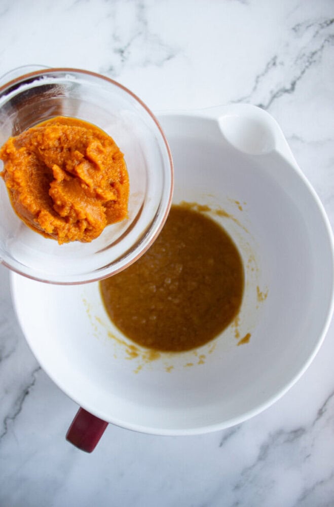 A glass bowl with pumpkin puree is being held over a white mixing bowl containing a brown liquid mixture on a marble countertop.
