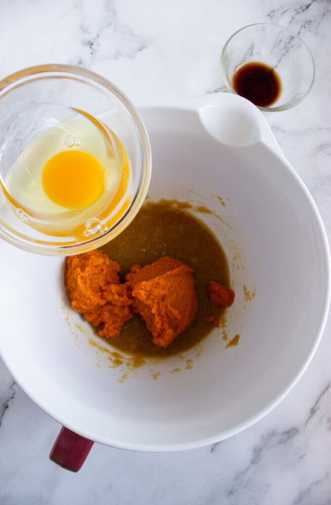 A mixing bowl with pumpkin puree and brown mixture, beside a bowl with an egg and oil, and a small bowl with vanilla extract, on a marble surface.