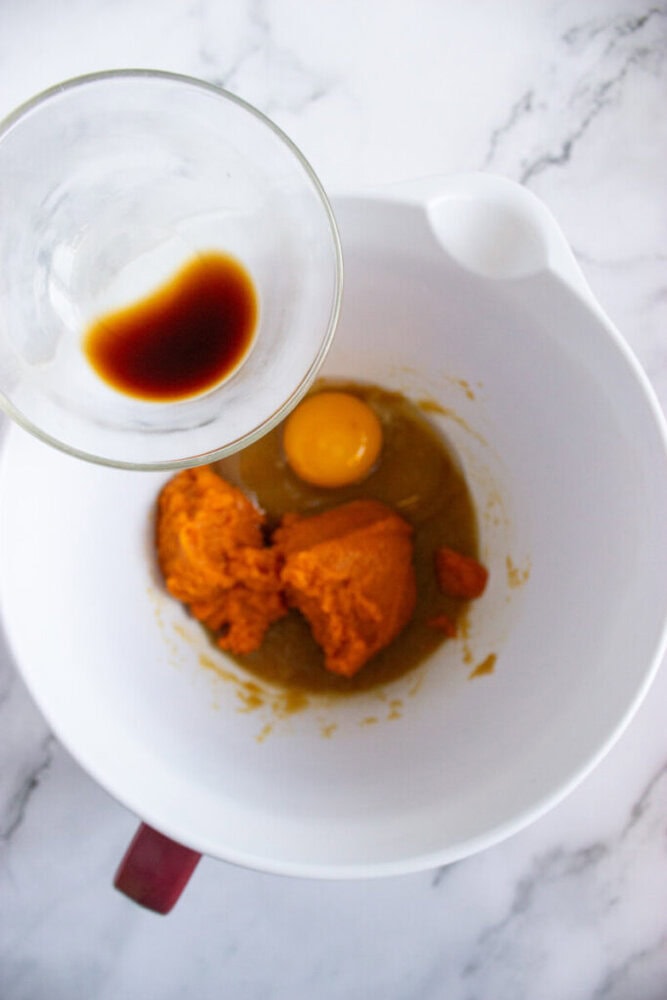 A mixing bowl with pumpkin puree, an egg, and sugar, with a small glass bowl of vanilla extract being held above it, ready to be added.