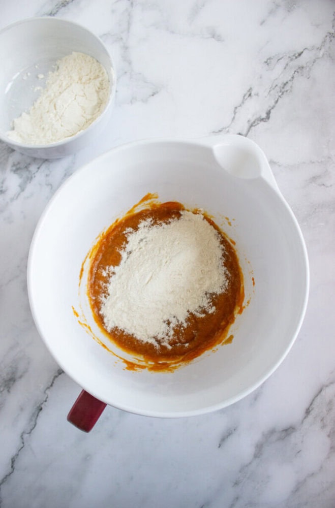 A white mixing bowl with orange batter and flour on top, next to a smaller bowl containing flour, on a marble countertop.