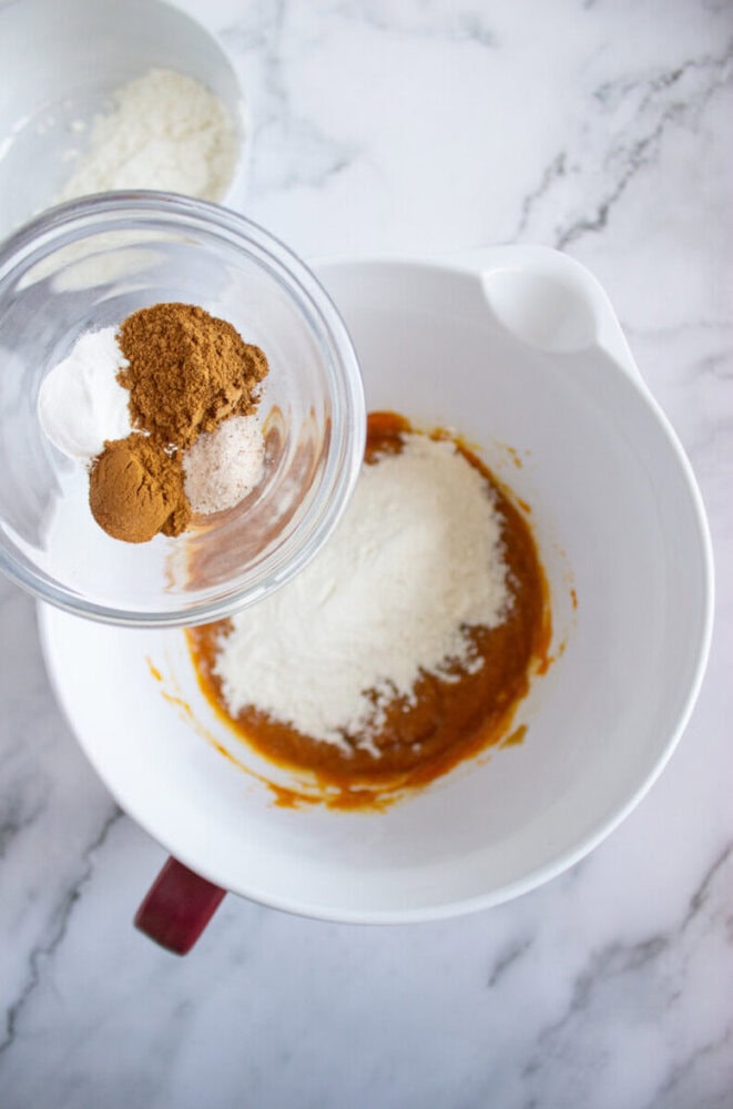 A hand holds a small bowl of spices above a larger mixing bowl containing flour and pumpkin puree on a marble countertop.