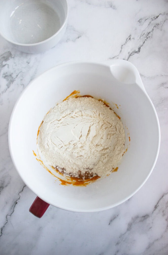 A white mixing bowl containing flour on top of wet ingredients sits on a marble countertop, with an empty bowl nearby.