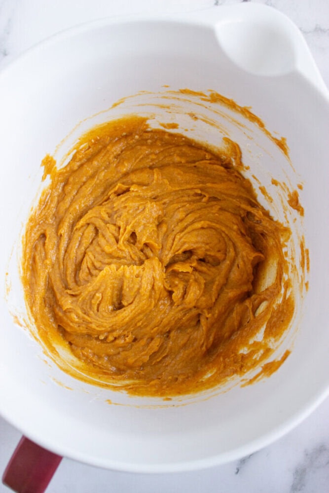 A white mixing bowl containing a thick, orange-colored batter being mixed, set on a white surface.