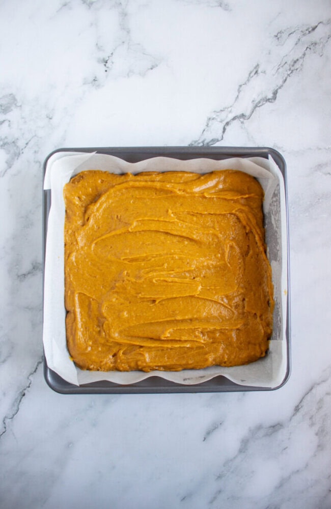 A square baking pan lined with parchment paper filled with an even layer of orange cake batter on a marble surface.