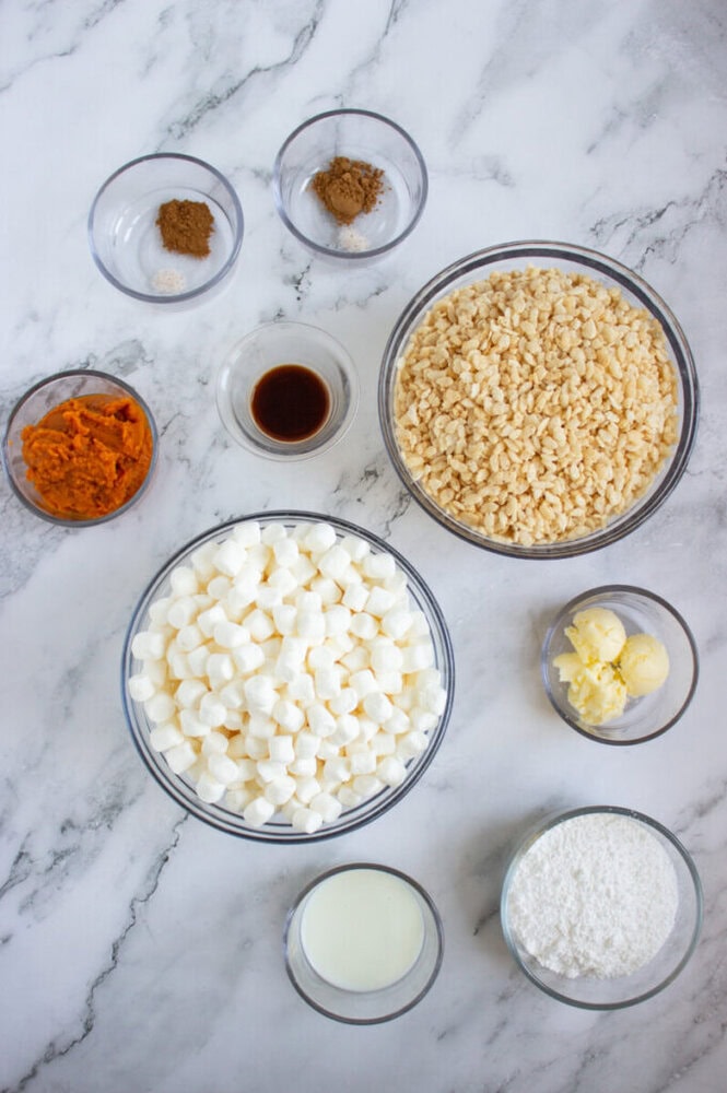 Bowls of Rice Krispies cereal, mini marshmallows, pumpkin puree, shredded coconut, butter, milk, vanilla extract, cinnamon, nutmeg, and salt arranged on a marble surface.