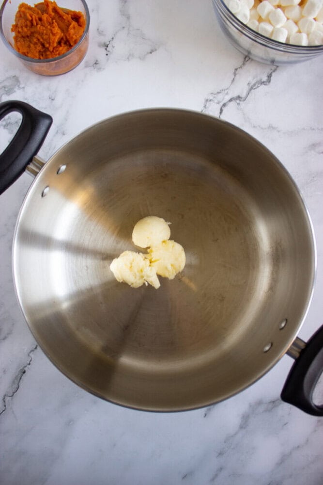 A stainless steel pot with scoops of butter inside sits on a marble countertop. Nearby are bowls containing marshmallows and orange puree.