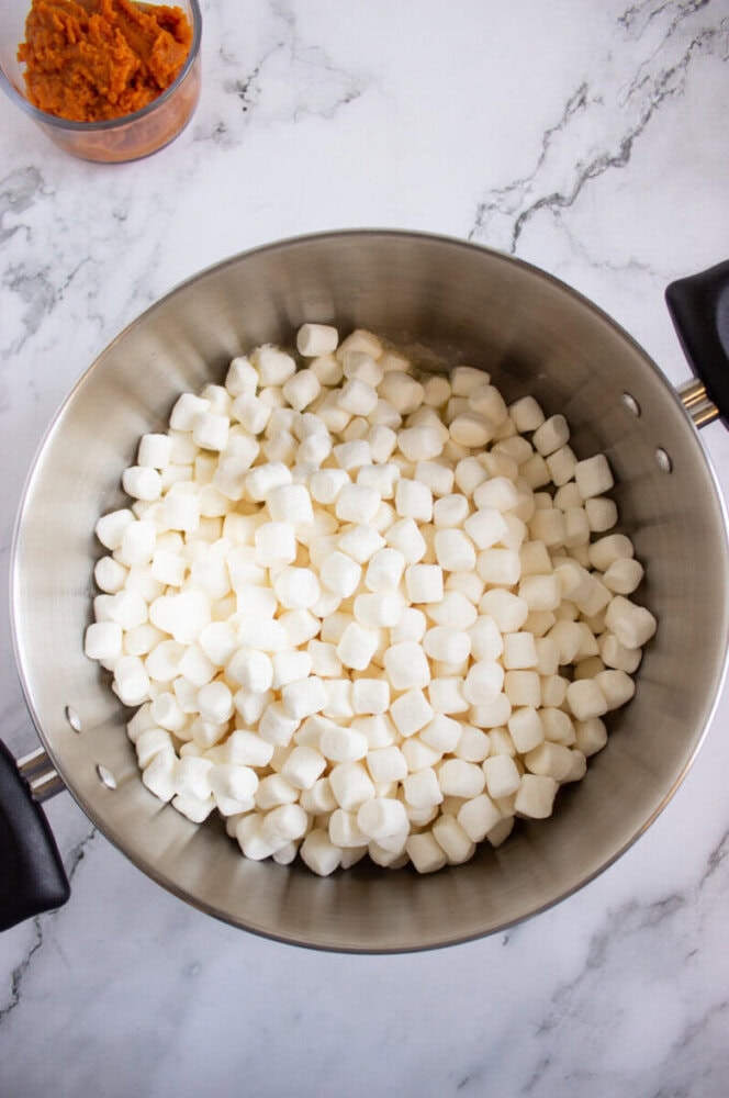 A metal pot filled with mini marshmallows sits on a marble surface; a small container of orange puree is partially visible in the corner.
