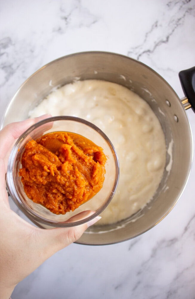 A hand holds a small bowl of pumpkin puree above a mixing bowl containing a creamy white mixture on a marble countertop.